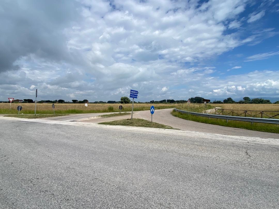 Straßenkreuzung inmitten von Feldern, mit Richtungsschildern und Straßenschildern unter einem teils bewölkten Himmel.