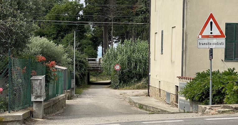 Narrow street between houses with a cyclist warning sign and a railway underpass at the end, surrounded by vegetation.