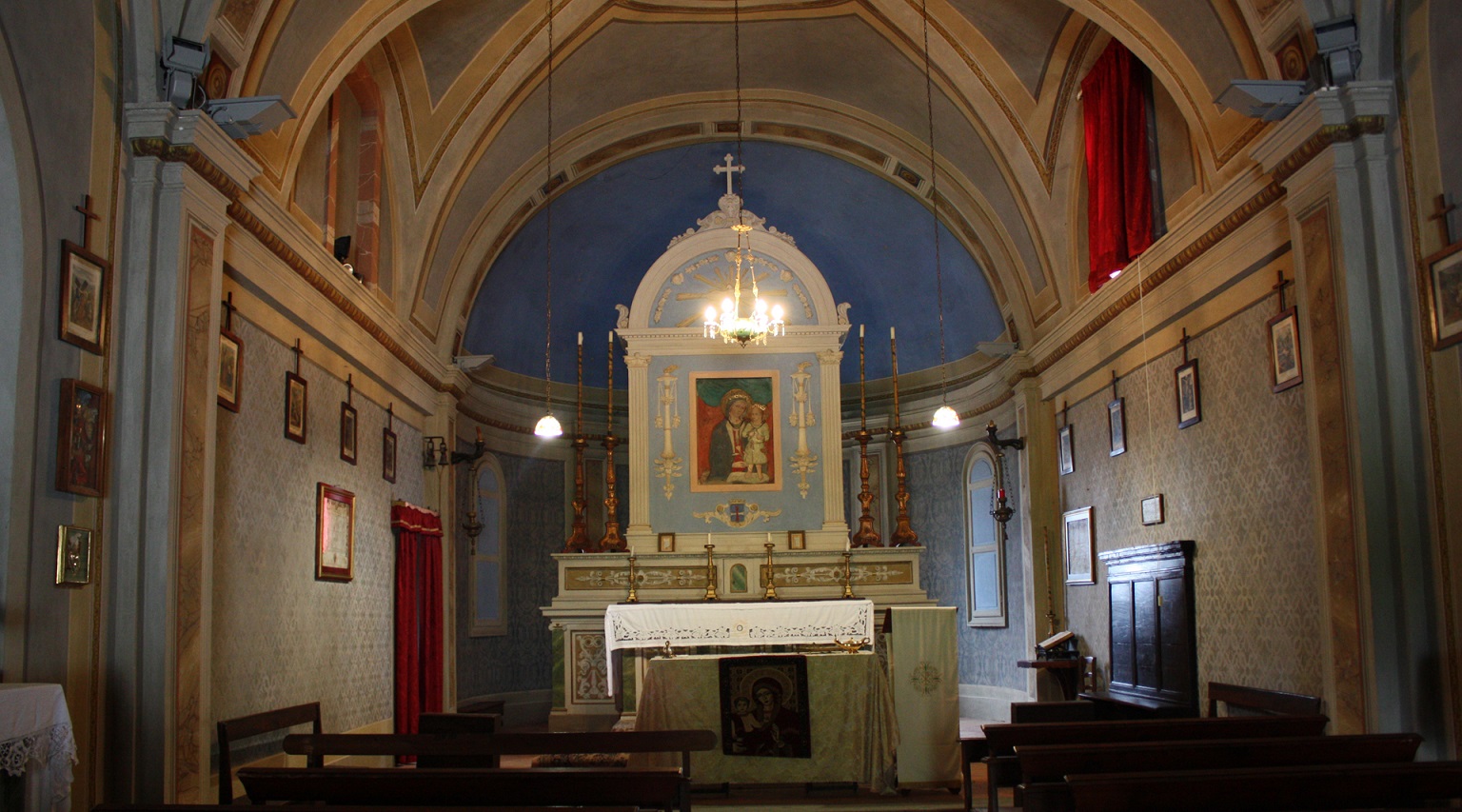 Interior of a church with a vaulted ceiling, central altar adorned with a painting of the Madonna and Child, and wooden pews.