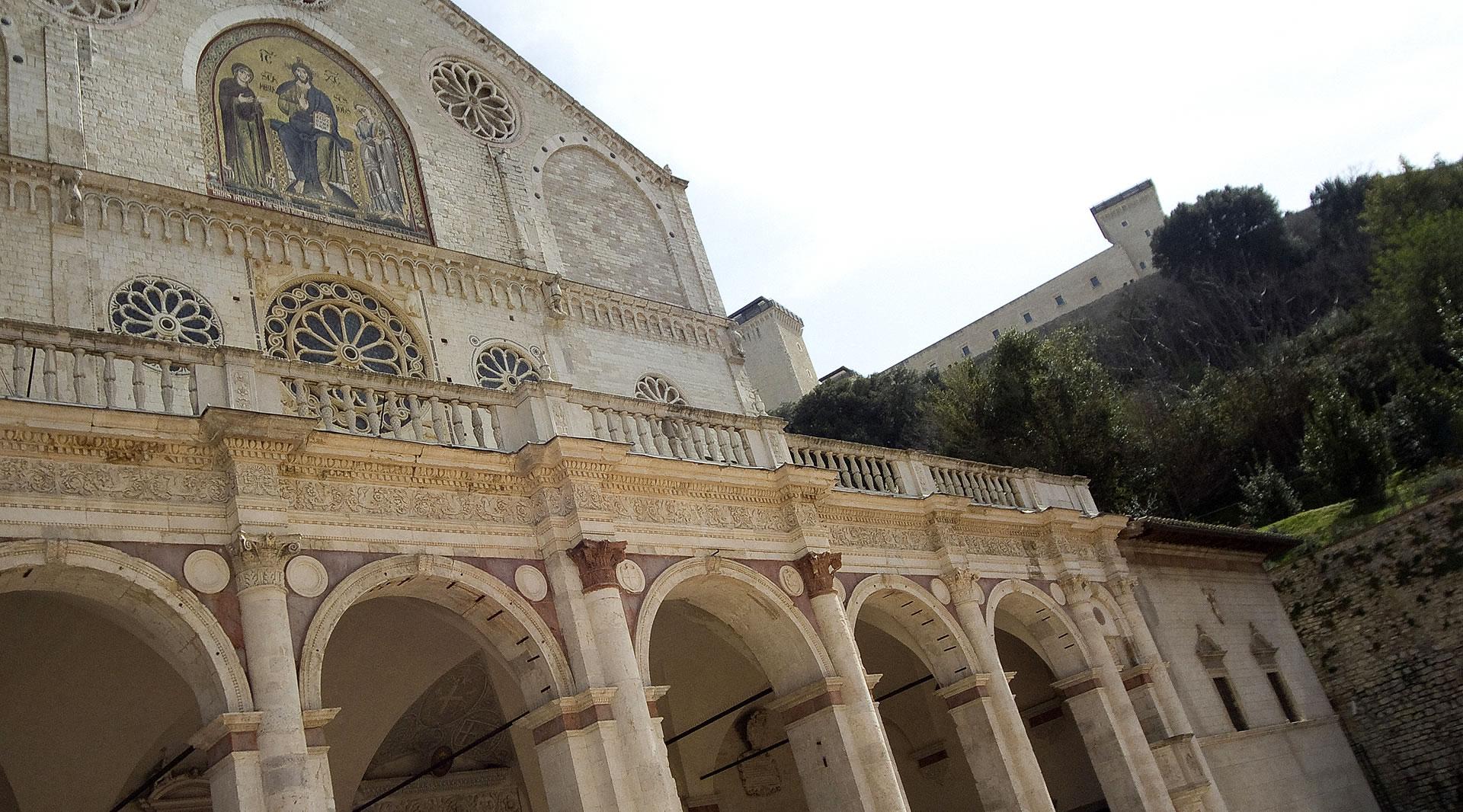 Façade of Spoleto Cathedral with arched portico, rose windows, golden mosaic, and the Rocca Albornoziana in the background.
