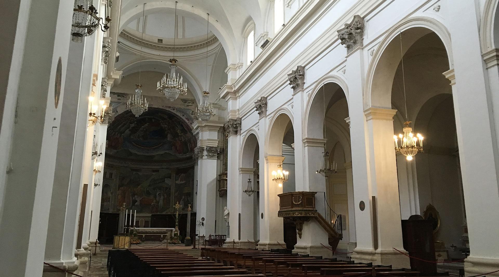 Interior of Spoleto Cathedral with a white nave, chandelier candelabras, wooden pulpit, and frescoes in the apse.