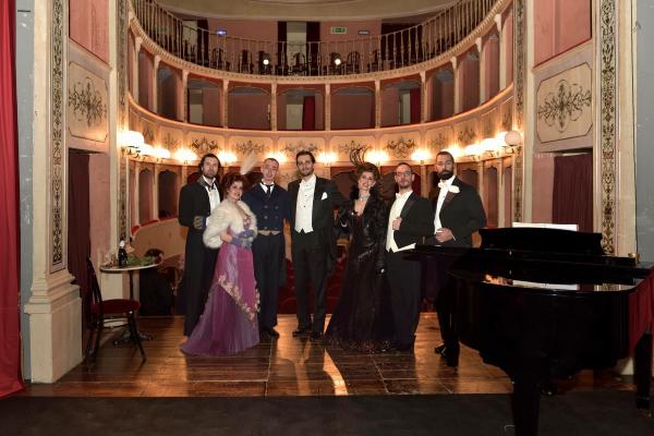  Blick von der Bühne des Teatro Cesare Caporali, mit Blick auf den Zuschauerraum und die eiserne Galerie. Der Saal, vollständig aus Holz, ist mit heller Farbe gestrichen und mit eleganten goldenen Stuckverzierungen versehen. Im Vordergrund stehen Künstler auf der Bühne. 