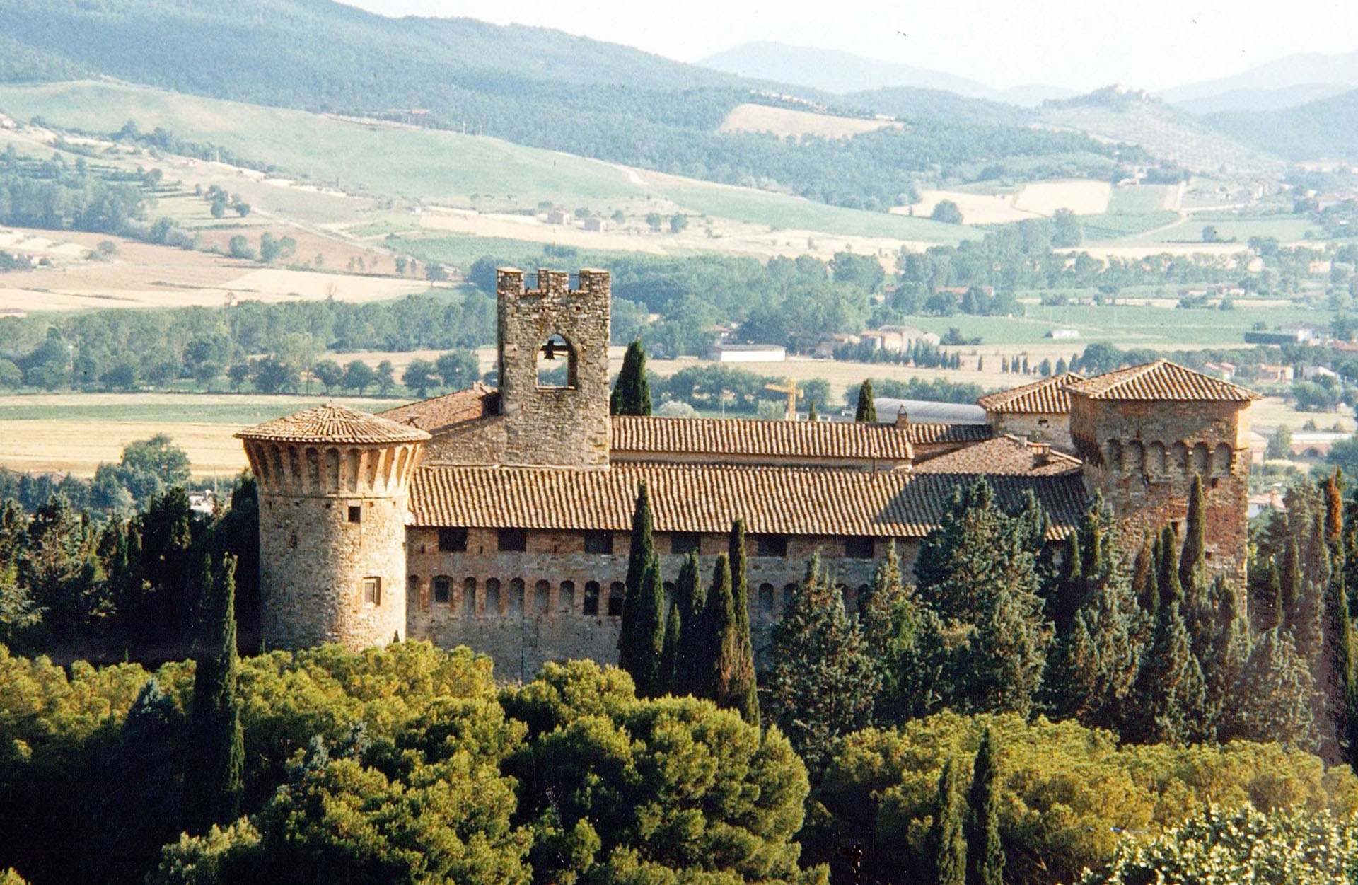 Castello medievale immerso nel verde delle colline umbre, con torri merlate e un paesaggio agricolo sullo sfondo.