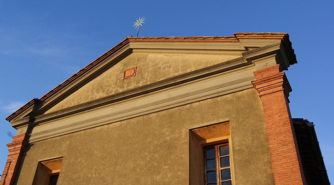 Fassade einer gelb verputzten Kirche mit Satteldach, roten Backsteinecken und einem goldenen Stern an der Spitze.