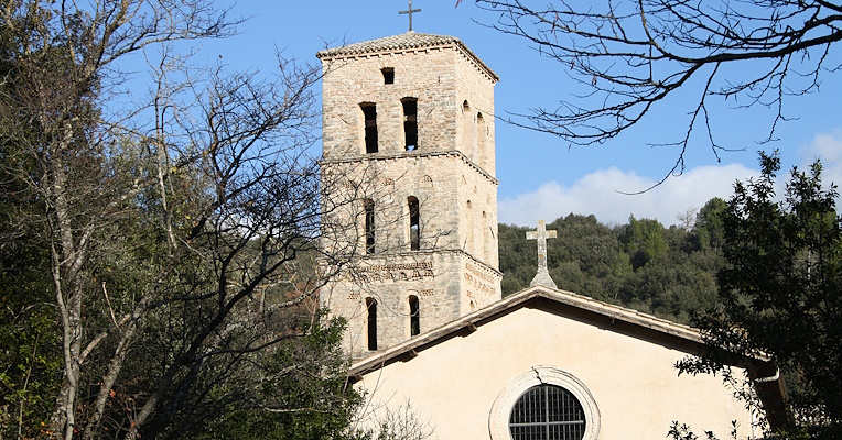 Fassade und Glockenturm der Abtei San Pietro in Valle in Ferentillo, mit einem Hintergrund aus Bäumen und blauem Himmel. Die Fassade hat ein großes rundes Rosettenfenster in der Mitte, während der Glockenturm mit quadratischem Grundriss und Rundbogenfenstern dahinter aufragt.