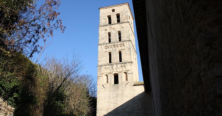 Campanile dell'Abbazia di San Pietro in Valle a Ferentillo visto dal basso, con alberi e cielo azzurro sullo sfondo. Il campanile, a pianta quadrangolare, è suddiviso in cinque piani con finestre ad arco e dettagli in laterizio.