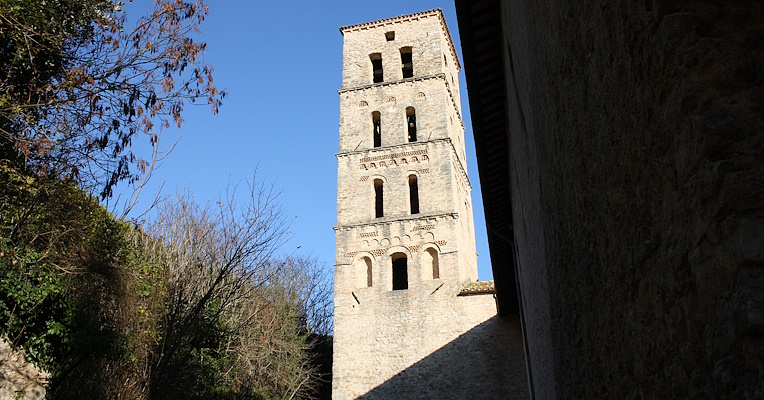 Glockenturm der Abtei San Pietro in Valle in Ferentillo von unten gesehen, mit Bäumen und blauem Himmel im Hintergrund. Der Glockenturm mit quadratischem Grundriss ist in fünf Stockwerke unterteilt und hat Rundbogenfenster und Ziegelsteindetails.