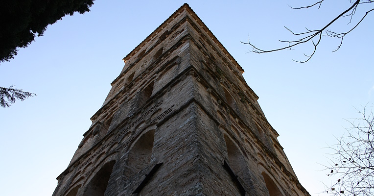 Ansicht von unten des Glockenturms der Abtei San Pietro in Valle in Ferentillo, mit einem hellblauen Himmel im Hintergrund. Der quadratische Glockenturm zeigt die Details der Rundbogenfenster und der Ziegelgesimse.