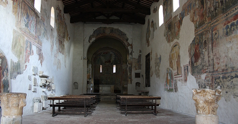 Interno dell'Abbazia di San Pietro in Valle a Ferentillo, con vista sulla navata centrale e sull'abside. Le pareti presentano affreschi medievali e finestre ad arco, mentre la navata è fiancheggiata da panche in legno e il soffitto è costituito da travi in legno a vista.