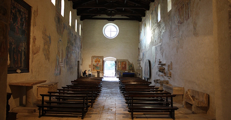 Innenansicht der Abtei mit Blick zum Eingang und dem runden Rosettenfenster über dem Eingang. Das Mittelschiff ist von Holzbänken flankiert, die zum Altar hin ausgerichtet sind, während die Wände mit Fresken dekoriert sind.