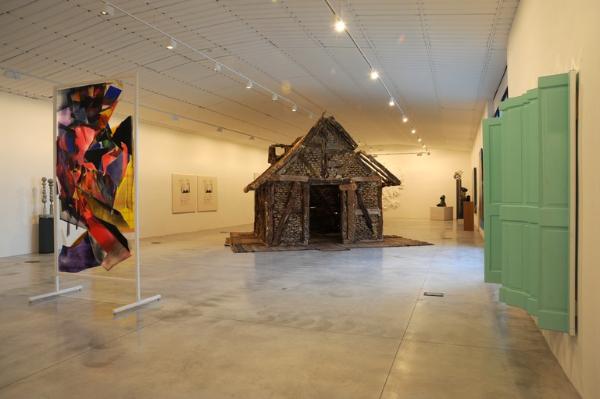  Une salle d'exposition avec une sculpture d'une cabane au centre, une œuvre abstraite à gauche et une fenêtre verte à droite. 