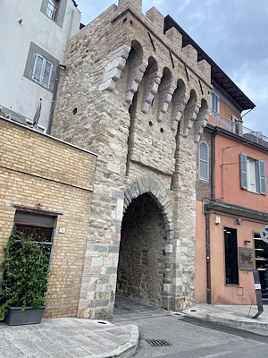 Ancient Porta Sant'Angelo in stone, with an arch and battlements, nestled among modern buildings in Bastia Umbra.