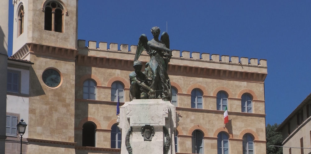 Commemorative bronze statue of a soldier and an angel in the centre of a square, with a historic building in the background.