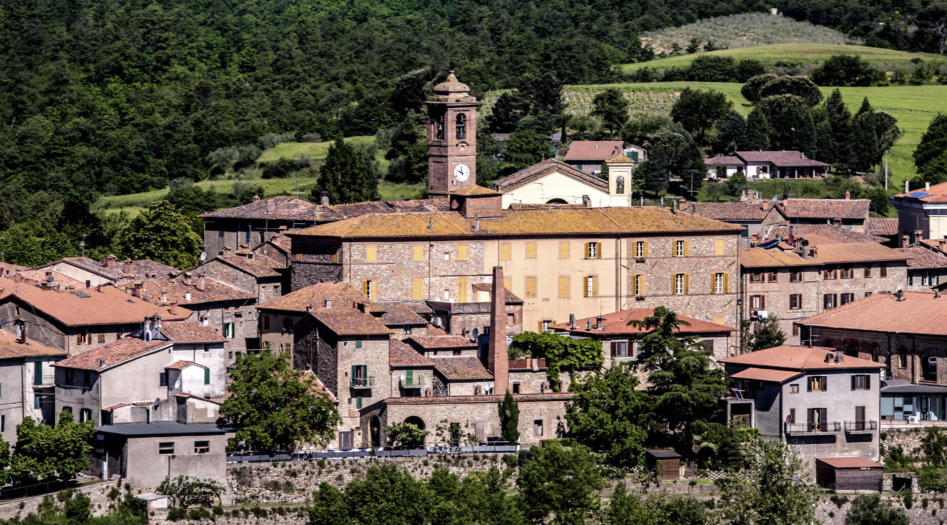 Village of Piegaro, nestled among green hills and trees, with stone buildings. In the foreground the chimney of the old glassworks