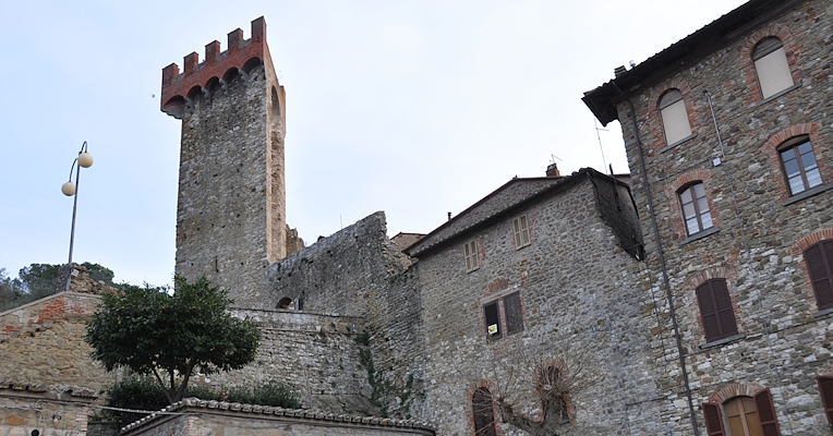 Torre medievale in pietra con merlature rosse, circondata da edifici in pietra con finestre ad arco e dettagli in mattoni.