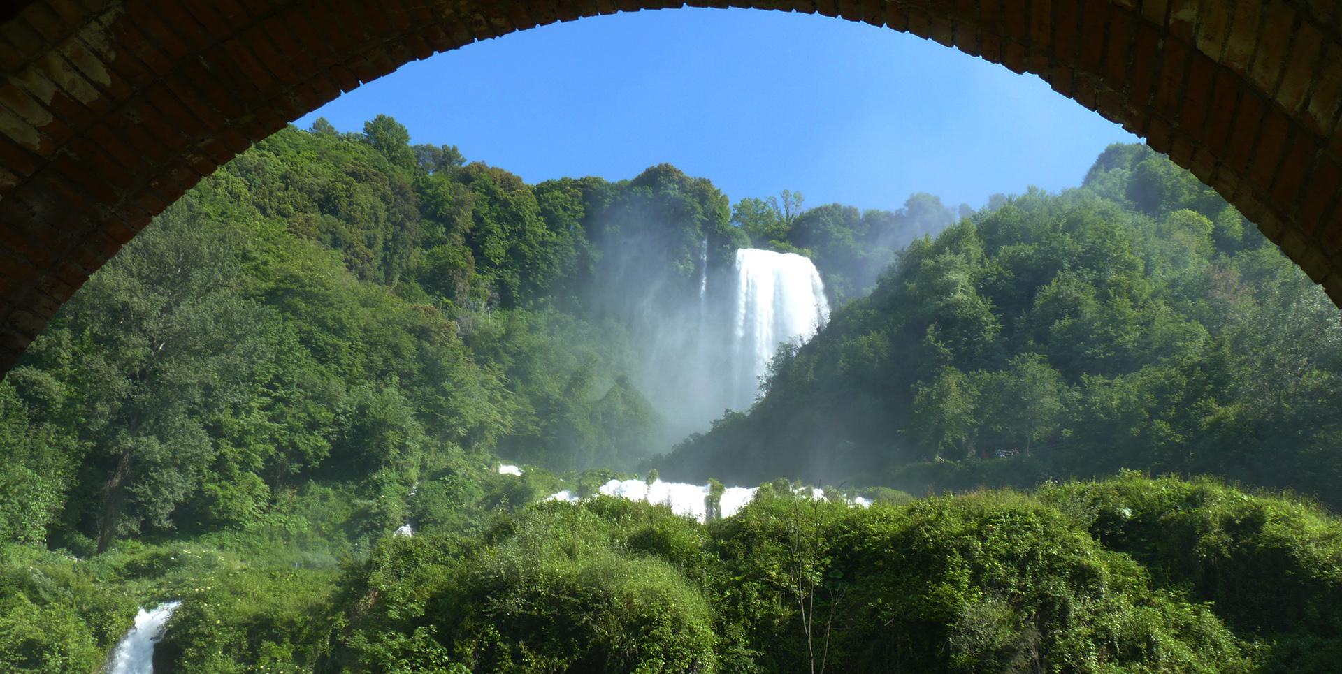 Vista della Cascata delle Marmore incorniciata da un arco di mattoni, immersa nella vegetazione con il cielo limpido sullo sfondo.
