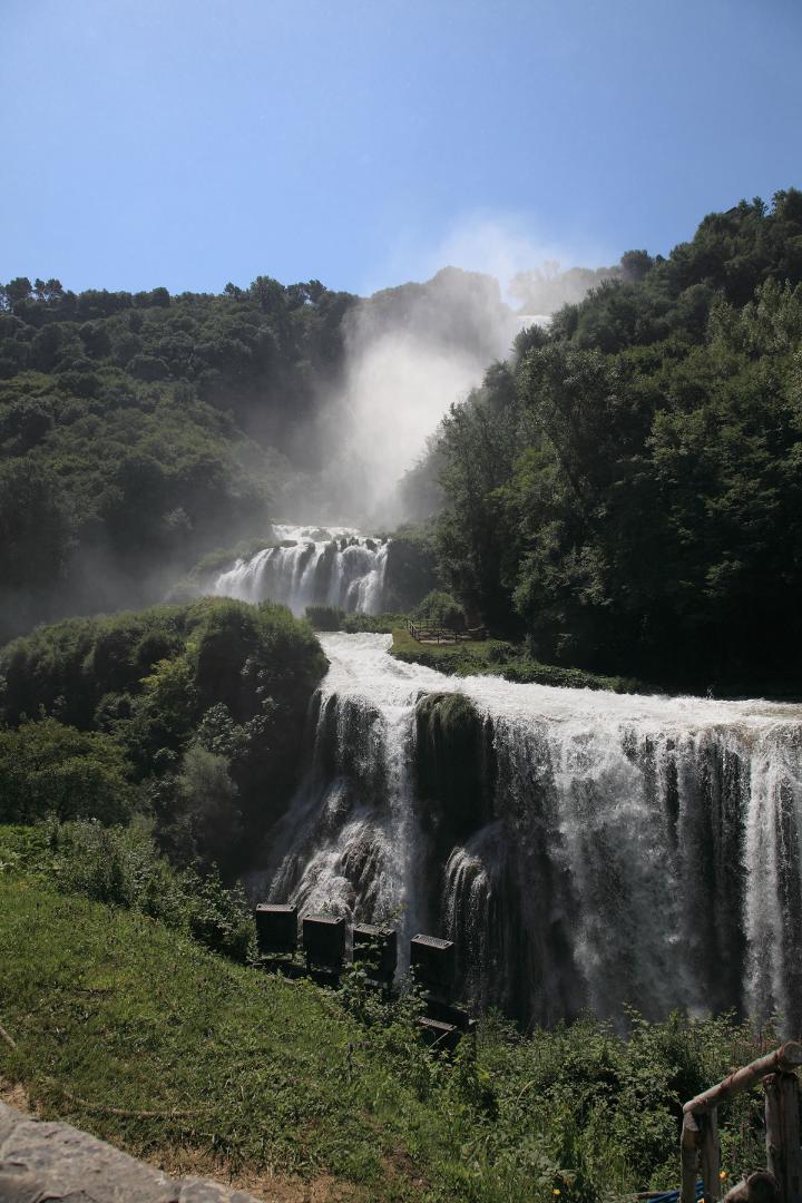 Cascata delle Marmore con i suoi salti d'acqua, incorniciata da rigogliosi alberi, sotto un cielo azzurro.