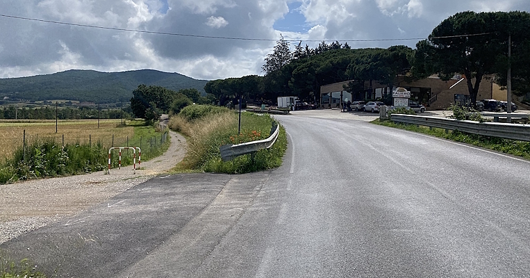 Route asphaltée longeant des champs et des collines, avec une piste cyclable à gauche et des bâtiments au loin.