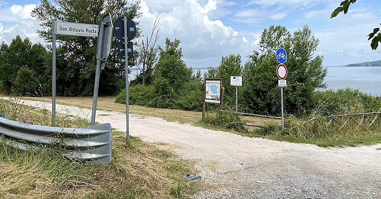 Sentiero sterrato con vista sul Lago Trasimeno, segnaletica ciclabile e indicazioni di accesso limitato in primo piano.