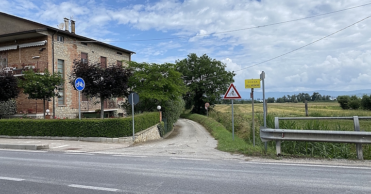 Route secondaire avec panneaux cyclables et avertissement de descente, bordée de champs et d'un bâtiment en briques.