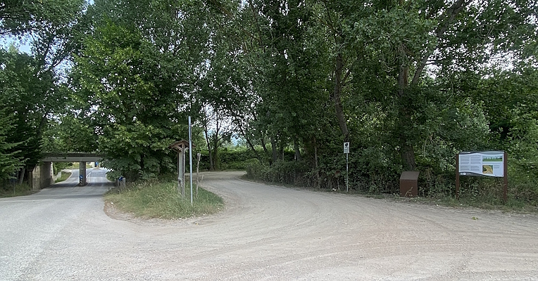 Junction on a dirt road, surrounded by trees, with informational signs and directions toward a bridge.