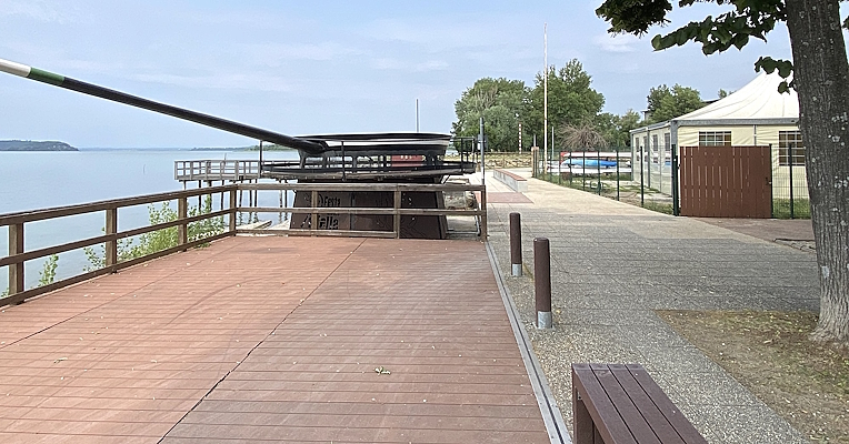 Passerella in legno lungo il lago, con recinzioni, alberi e strutture sullo sfondo, situata vicino alla riva del Trasimeno.