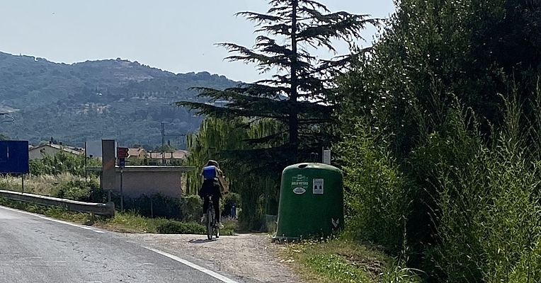 Radfahrer auf einer Straße in der Nähe eines Glasrecycling-Containers, umgeben von Vegetation und Hügeln im Hintergrund.