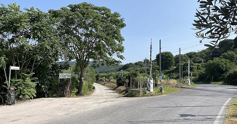 Junction on a paved road with access to a campsite and a level crossing, immersed in a hilly green area.