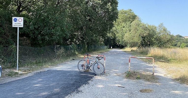 Schotterweg mit rot-weißen Schranken, einem Schild für das Durchfahrtsverbot und einem daneben abgestellten Fahrrad.