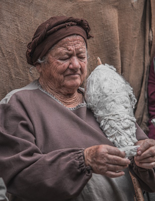 Immagine di una signora che tiene una rocca con dei ciuffi di lana tra le braccia mentre lavora un ciuffo di lana ancora da filare Credits photo: Francesco Andreoli