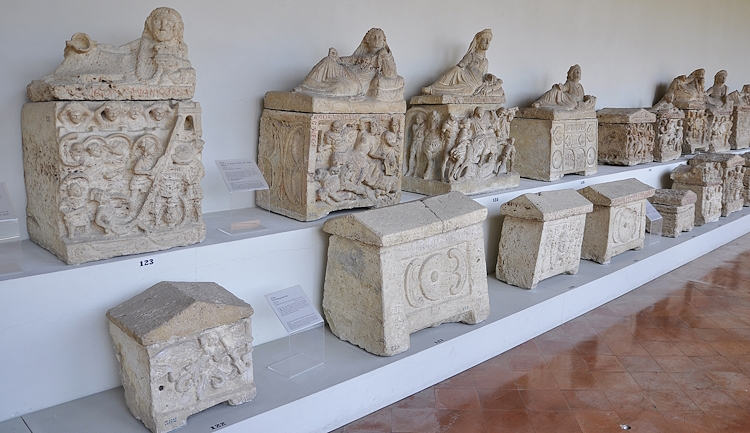 Urnes cinéraires en calcaire blanc, dont certaines avec un toit représentant le défunt en position semi-allongée, au Musée archéologique national de l’Ombrie.