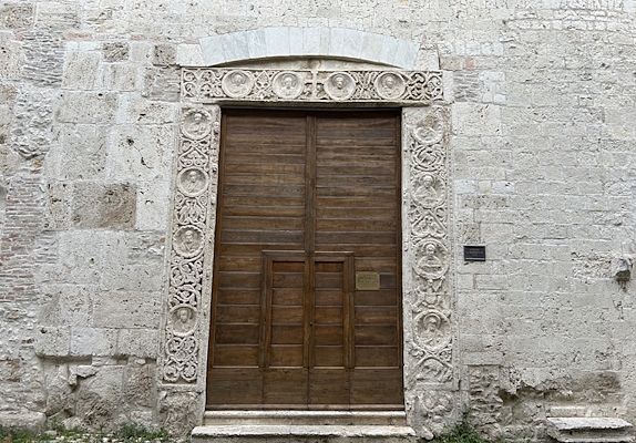 Wooden door of the former Church of San Domenico in Narni, now Bortolotti Auditorium, adorned with a marble frame featuring a series of medallions carved with portraits of religious figures. Above the door, a white stone arch completes the monumental entrance.