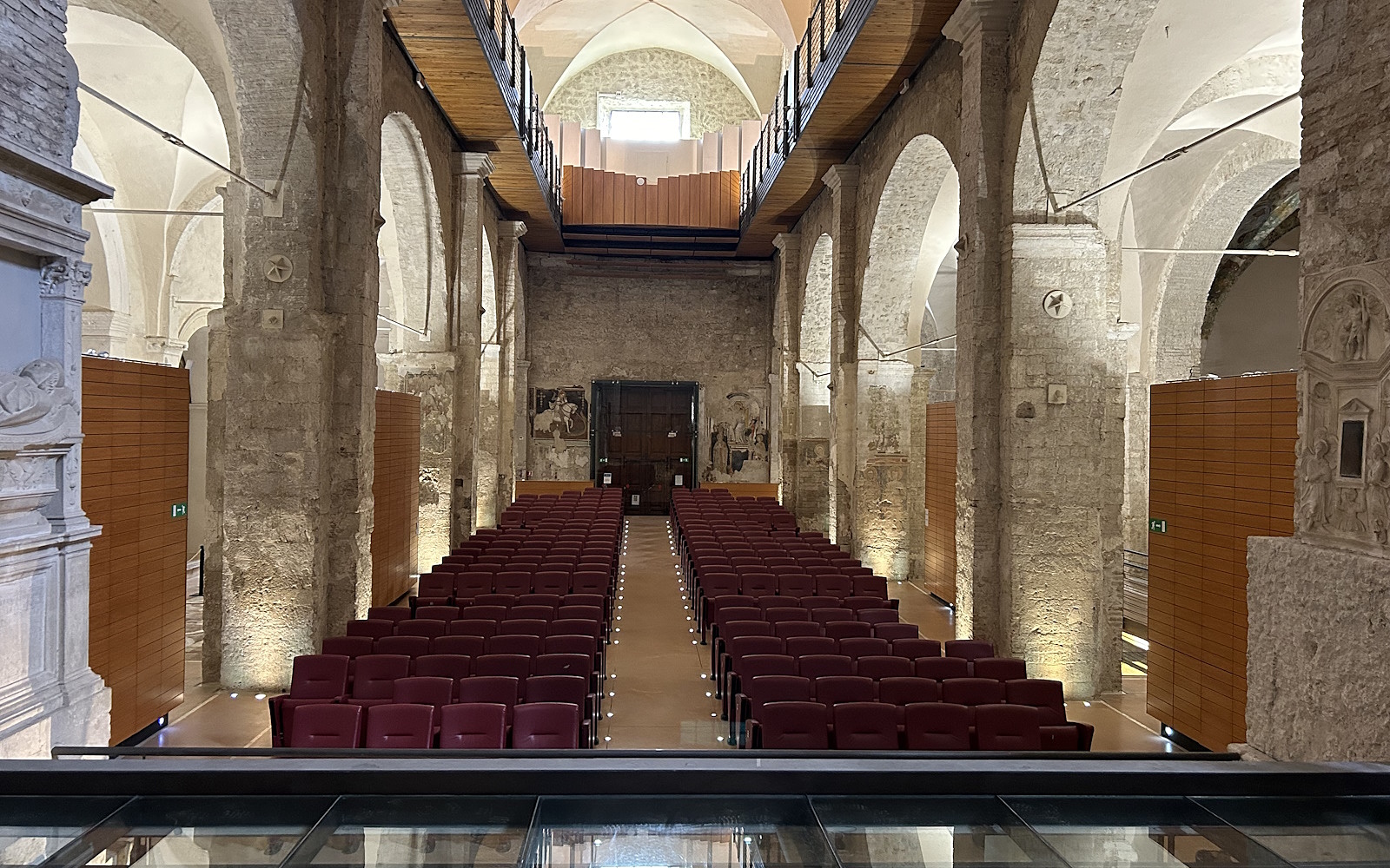 Interior of the Bortolotti Auditorium in Narni with rows of red seats arranged symmetrically, stone walls, a vaulted ceiling with ribbed vaults, and a wooden walkway on the upper floor that runs along both sides of the hall. In the background, a large wooden door and partially visible frescoes adorn the wall.