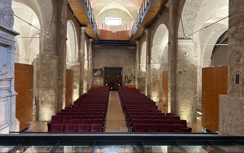  Interior of the Bortolotti Auditorium in Narni with rows of red seats arranged symmetrically, stone walls, a vaulted ceiling with ribbed vaults, and a wooden walkway on the upper floor that runs along both sides of the hall. In the background, a large wooden door and partially visible frescoes adorn the wall. 