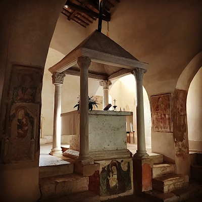 Autel en travertin, surmonté d'un ciborium avec colonnes et un toit à quatre pans. Les murs du bâtiment sont décorés de fresques religieuses représentant des figures sacrées, visibles sur les côtés de l'autel et les murs environnants.