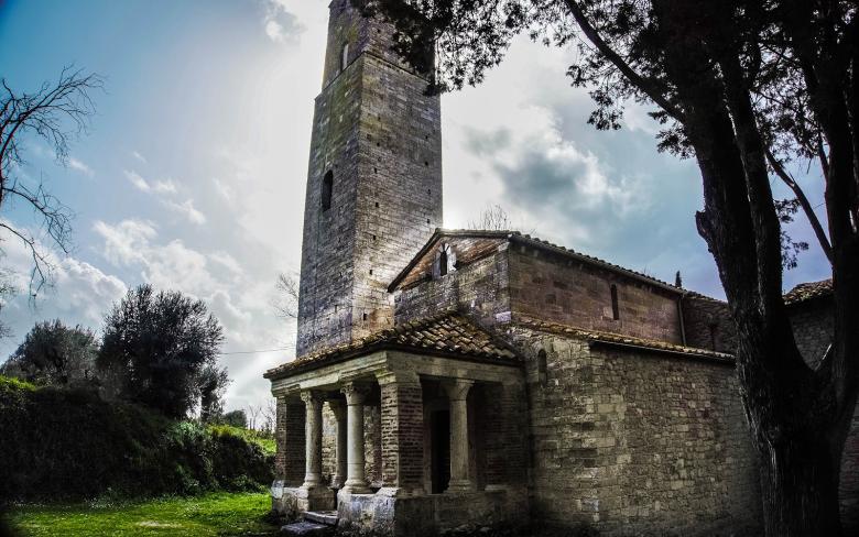  Kirche Santa Pudenziana mit einem steinernen Glockenturm, einem Portikus mit Säulen und einem Ziegeldach. Im Hintergrund ein teilweise bewölkter Himmel und Bäume. 