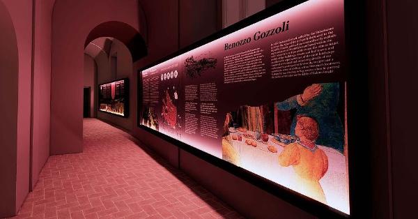  A museum corridor with walls and arches illuminated by a dark pink light. On the right, a long informational panel features texts and images dedicated to Benozzo Gozzoli. 