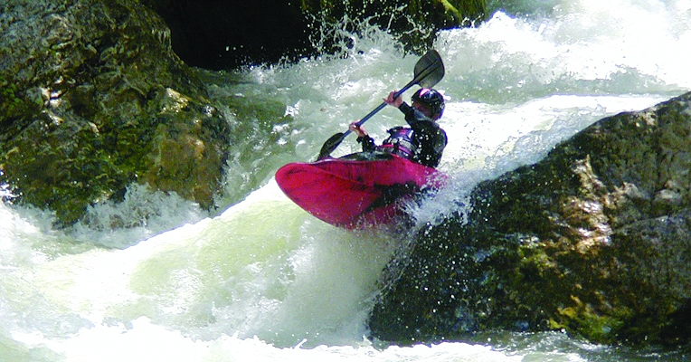 Red kayak led by a man holding a paddle while jumping a rapid on a river course