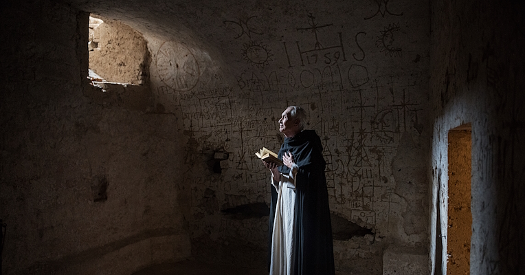 Homme en habits historiques debout avec un livre à la main dans la cellule de Giuseppe Andrea Lombardini, avec des murs couverts de graffitis et de symboles ésotériques. La lumière entre par une petite fenêtre, illuminant partiellement la pièce.