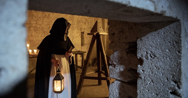 Homme en habits historiques tenant une lanterne dans la Chambre des Tourments, éclairée par des bougies. À l'arrière-plan, on peut apercevoir des instruments de torture.