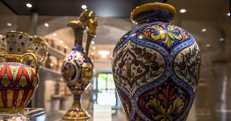 Lustreware majolica vases on display in the Rubboli Workshop Museum. In the foreground, a large vase decorated with intricate floral and geometric motifs in bright blue, yellow, red and green. In the background, other vases and ceramic objects, including an amphora with an elaborate handle and a jug, also richly decorated with similar motifs.