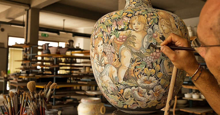 A local craftsman from Gubbio decorating a large historiated majolica vase. The vase features an elaborate floral design and figures of cherubs. In the background are shelves full of other ceramics and work tools. On the table, next to the craftsman, are various brushes and tools needed for painting and decorating the majolica.