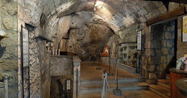 Medieval kiln located in the Pozzo della Cava archaeological complex in Orvieto. The scene is characterised by stone walls and ceilings, with clearly visible arched architecture. There are several staircases leading to lower levels, while various ceramic artefacts and other historical artefacts are displayed along the walls. The exhibits are protected by ropes and poles, creating a guided tour for visitors.