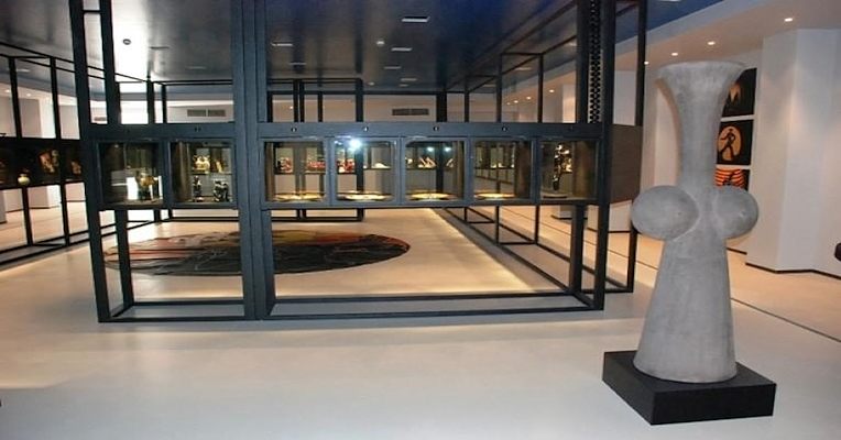 Entrance to the Rometti Gallery in Umbertide. The exhibition space is characterised by a modern, minimalist design with a white floor and white walls. In the centre of the room is a large black metal structure containing several illuminated showcases with ceramic objects. On the right side of the picture, there is a tall grey ceramic sculpture on a black pedestal, characterised by a tapered shape with two prominent spherical elements. In the background, other works of art are displayed along the walls.