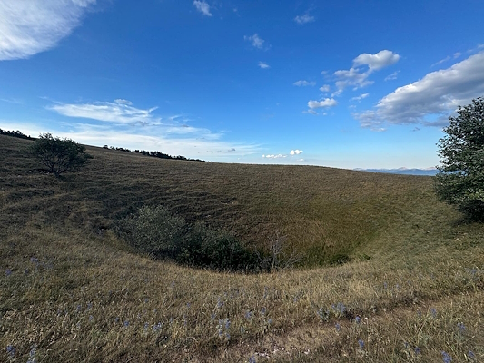 A panoramic view of a hilly landscape with a deep sinkhole. Shrubs and trees surround the edges of the depression, while the blue sky is interspersed with a few white clouds.