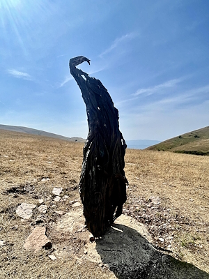 Eine dunkle, verbogene Holzskulptur unter einem blauen, hügeligen Himmel.