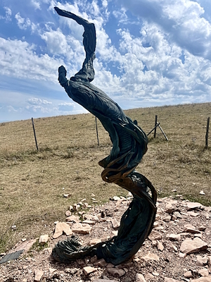 Sinuous, twisting sculpture, dark green with bronze highlights, on rocky ground with cloudy sky and hills in the background.