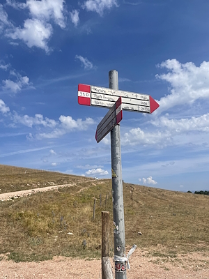 Palo con segnaletica escursionistica, colline e cielo limpido sullo sfondo.