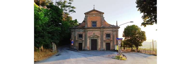 Immagine: Una piccola chiesa in mattoni, circondata da alberi, si trova a un bivio con cartelli stradali. 