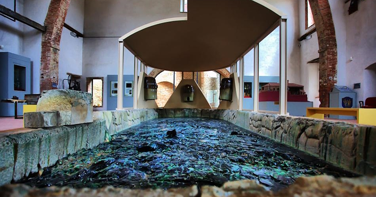  Interior of the Glass Museum in Piegaro, with a stone basin containing fragments of colored glass and exposed brick arches. 